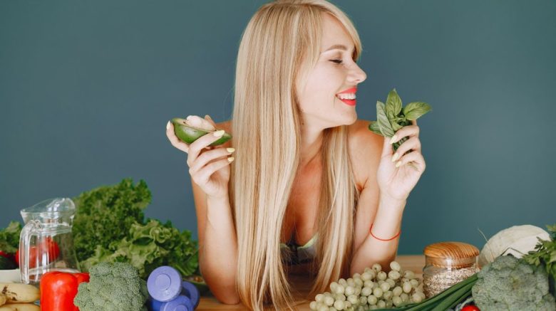 A smiling woman holding avocado and basil, surrounded by fresh vegetables and fruits for healthy eating.