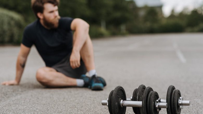 Serious bearded male in activewear resting after tiring workout on asphalt road with heavy dumbbells in soft focus